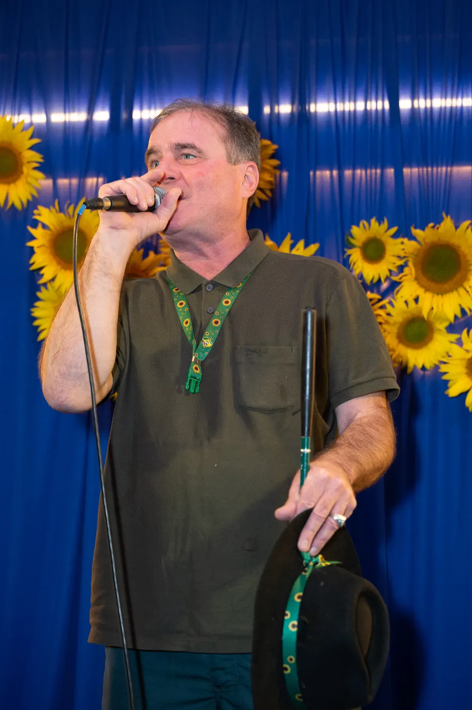 Zezinho Fisioterapeuta ao microfone durante um ato de pré-campanha, com painel de girassóis ao fundo, segurando o chapéu preto na outra mão.