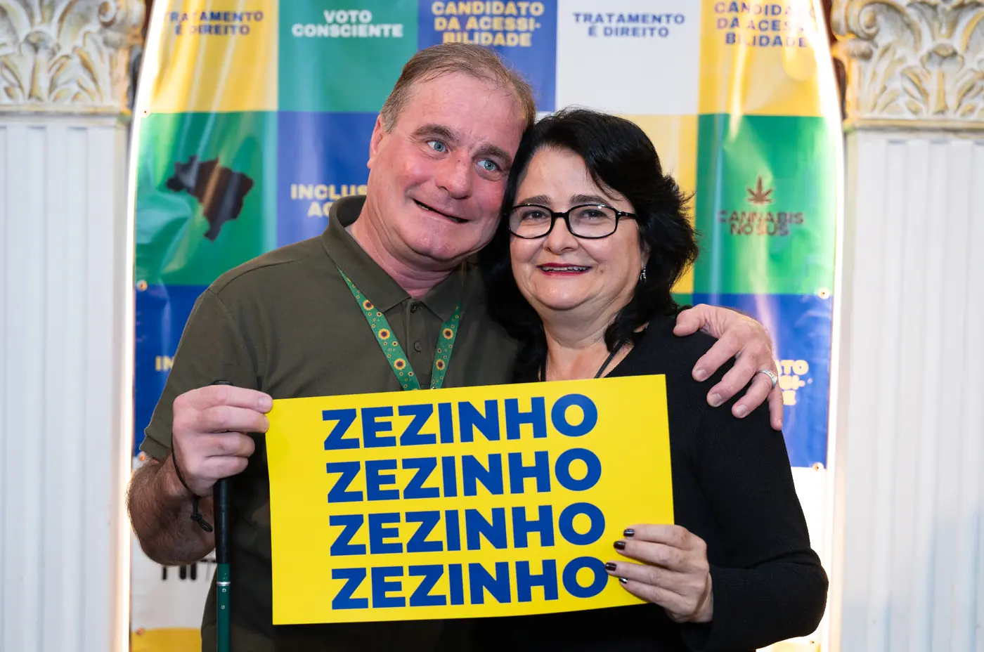 Zezinho abraçado à esposa, ela segurando um cartaz amarelo com o nome ZEZINHO em letras azuis, em ato de pré-campanha.