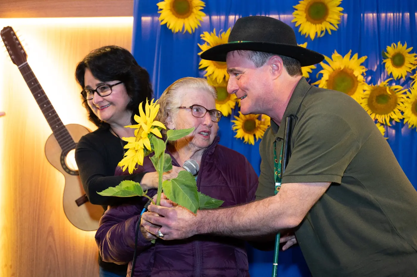 Zezinho, de chapéu preto, presenteando sua mãe com um girassol amarelo durante evento de pré-campanha, com painel de girassóis ao fundo.